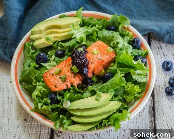 Superfood Salmon with Balsamic Blueberry Sauce is full of no-guilt delicious greens, avocados and a delicious piece of salmon A white bowl with an orange rim is filled with green lettuce that's adorned with a thick piece of salmon topped with a balsamic blueberry sauce. Sliced avocado sits on each side of the salad and fresh blueberries are mixed into the greens.