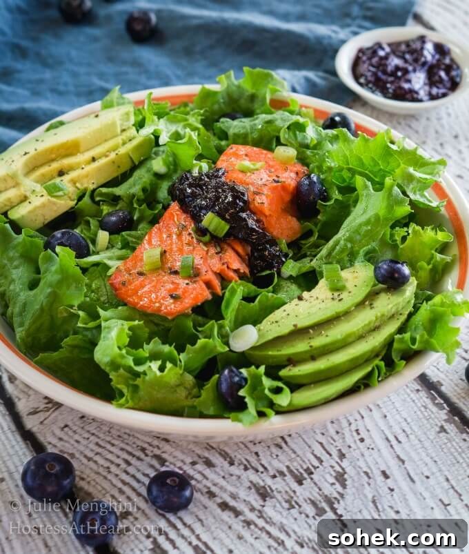 Beautiful bowl of Superfood Salmon Salad with Balsamic Blueberry Sauce is full of delicious healthy ingredients. It's a complete meal in a bowl that will keep you satisfied for hours. A white bowl with an orange rim is filled with green lettuce that's adorned with a thick piece of salmon topped with a balsamic blueberry sauce. Sliced avocado sits on each side of the salad and fresh blueberries are mixed into the greens over a white board.