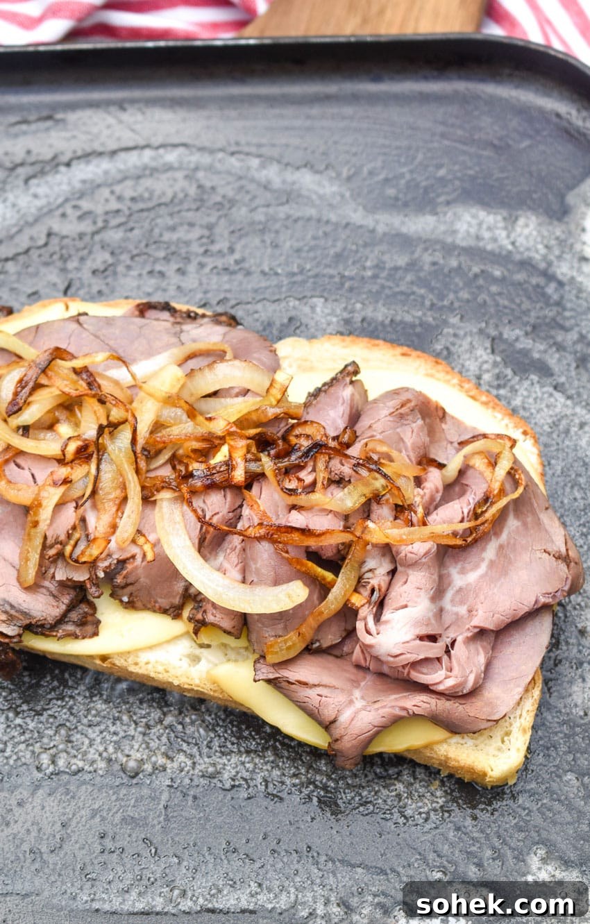a slice of sourdough bread topped with sliced gouda cheese, roast beef, and caramelized onions on a buttered skillet