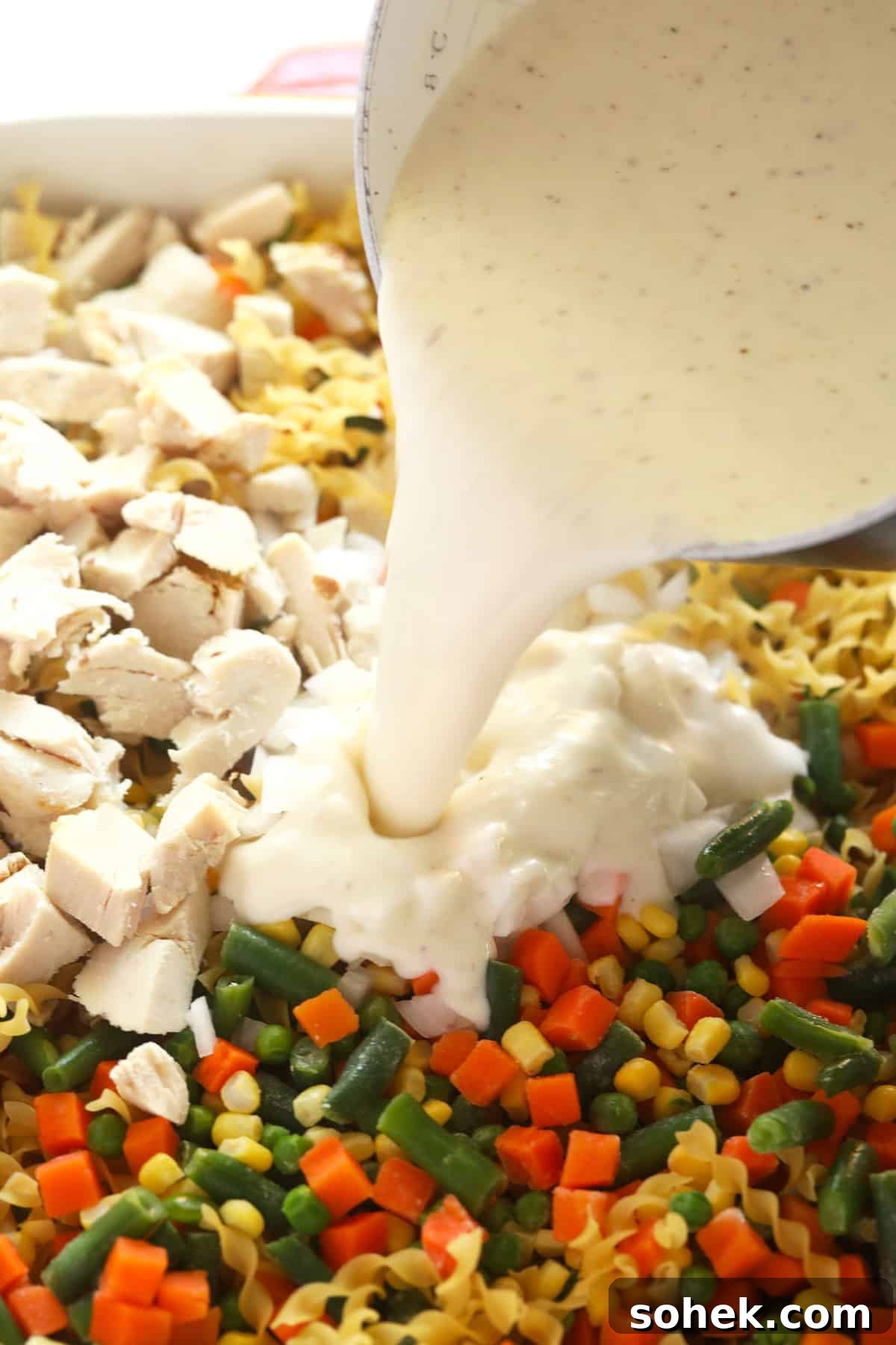 This Creamy Chicken Noodle Casserole is comfort food at its finest! Everything you love about chicken noodle soup, but as a flavor-packed casserole. Enjoy! Creamy cheese sauce being poured over a chicken noodle casserole before being baked.