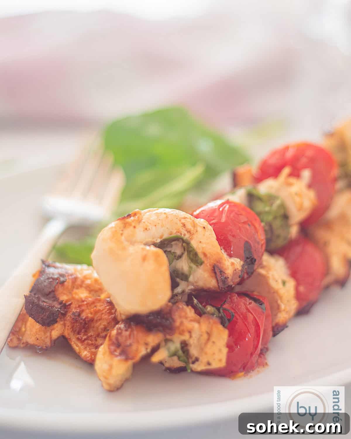 Mozzarella-Stuffed Grilled Chicken Skewers with Basil and Tomato 2 A photo of a white plate with three skewers of stuffed chicken with mozzarella and basil and grilled cherry tomatoes.