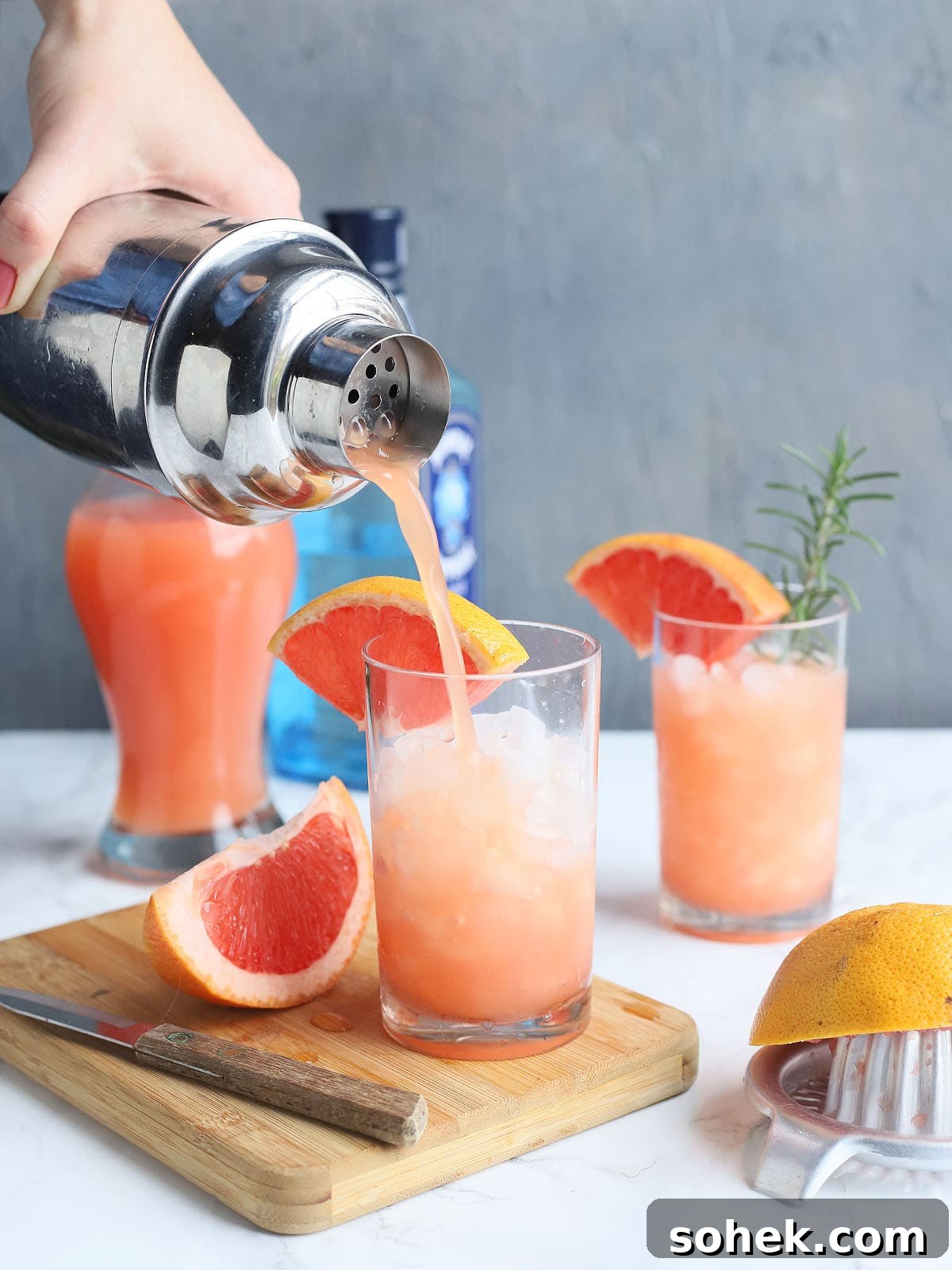 Rosemary Gin Grapefruit Cocktail Recipe with Citrus Twist 8 hand pouring a grapefruit cocktail from a shaker into a glass garnished with a grapefruit wedge and a sprig of rosemary