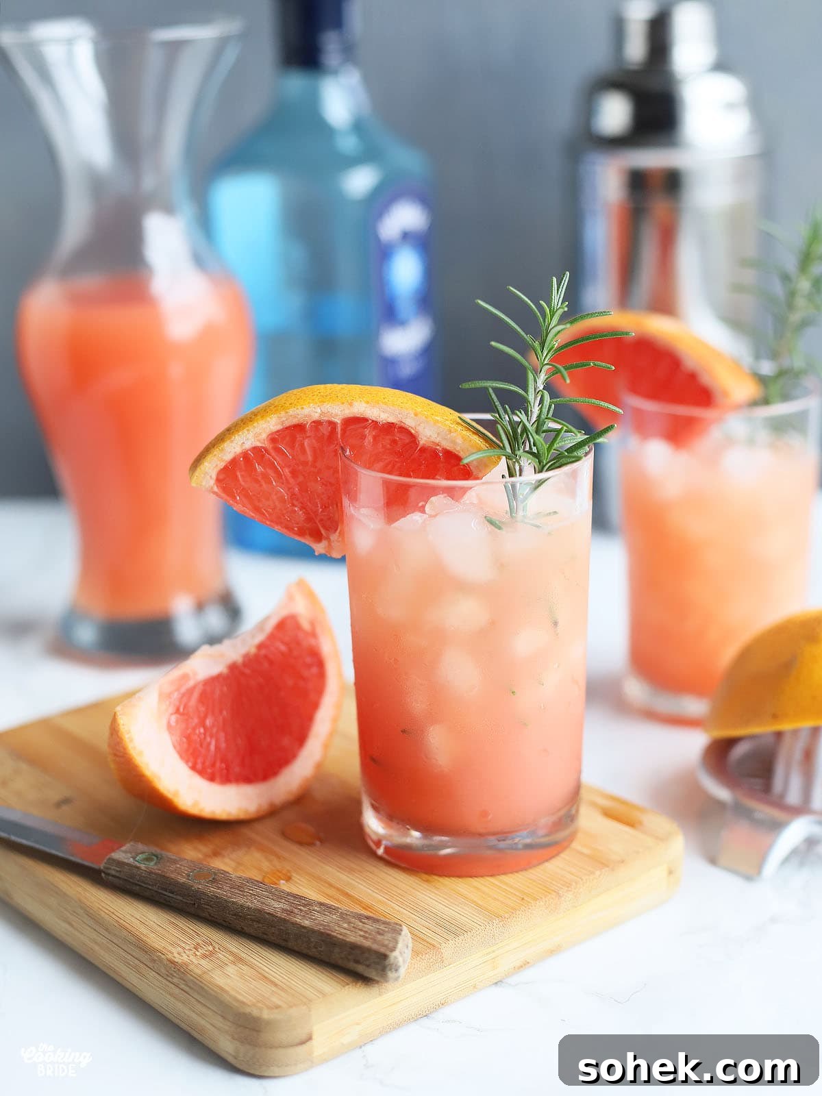 rosemary and gin grapefruit cocktail in a glass of ice garnished with a grapefruit wedge and a sprig of rosemary, slice of grapefruit and a knife to the side with another cocktail, grapefruit juice, bottle of gin and shaker in the background