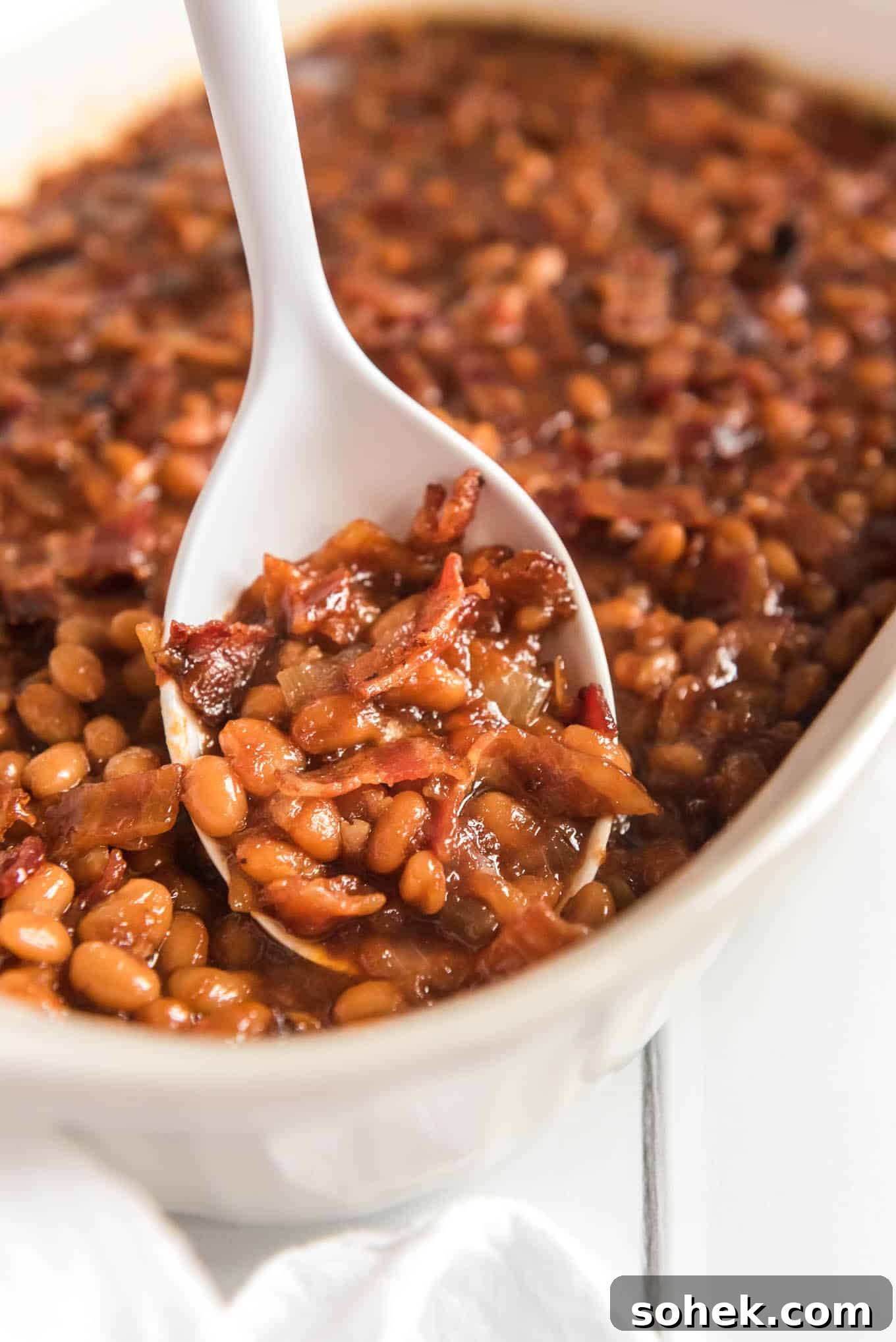 A white spoon scooping some bacon baked beans from the dish.