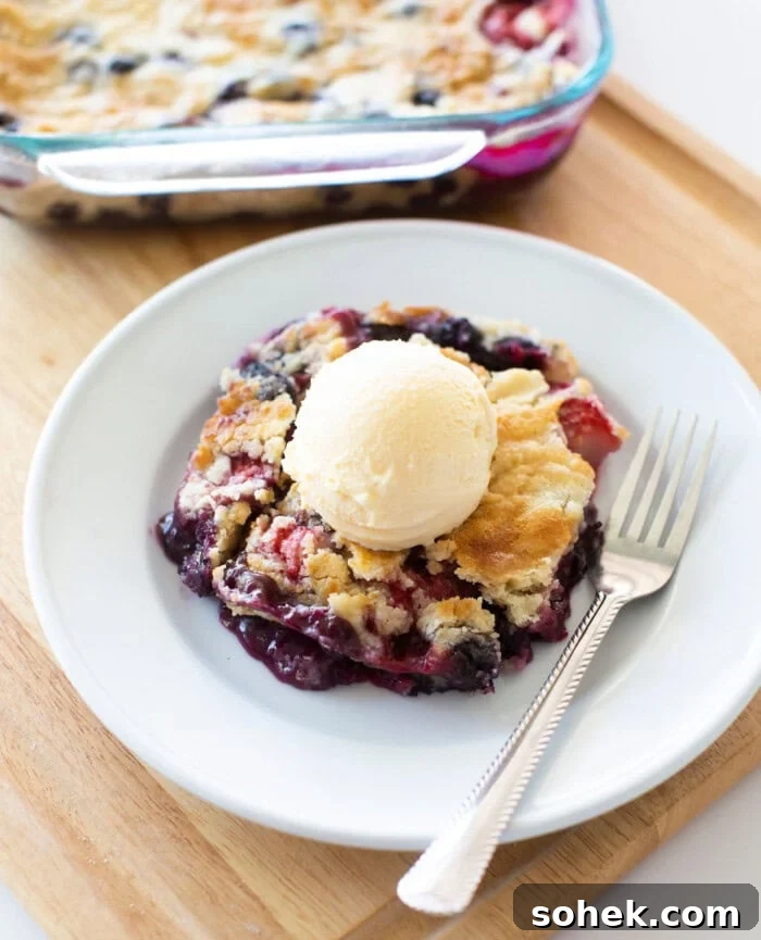 Viral Berry Dump Cake Recipe: Quick No-Fuss Dessert PMID 13 A slice of berry cobbler topped with a scoop of vanilla ice cream is served on a white plate with a fork. The baking dish with the rest of the cobbler is visible in the background on a wooden surface.