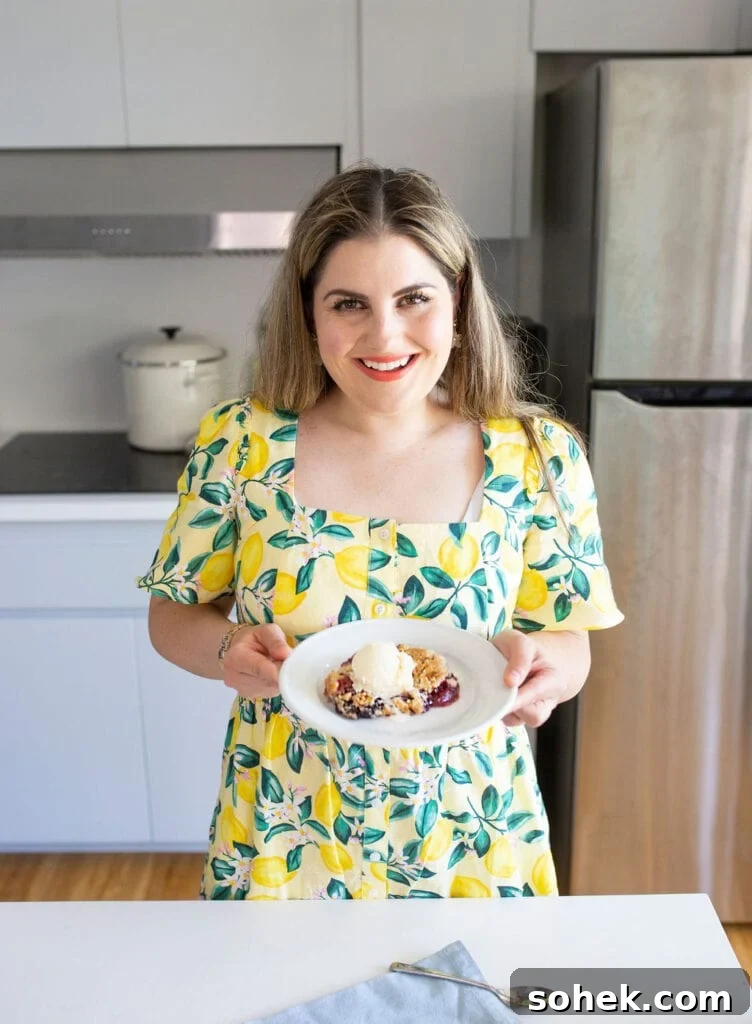 Viral Berry Dump Cake Recipe: Quick No-Fuss Dessert PMID 5 A woman in a yellow dress with a lemon pattern holds a white plate with a dessert. She stands in a kitchen with a stove, pot, and refrigerator in the background, smiling at the camera.