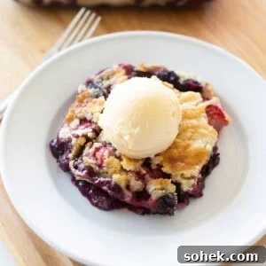 Viral Berry Dump Cake Recipe: Quick No-Fuss Dessert PMID 6 A serving of mixed berry crumble topped with a scoop of vanilla ice cream on a white plate. The dessert is set on a wooden surface with a fork and a hint of the larger dish in the background.