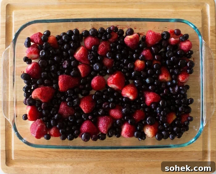 Viral Berry Dump Cake Recipe: Quick No-Fuss Dessert PMID 7 A glass baking dish filled with a mix of strawberries and blueberries, placed on a wooden surface. The fruit is vibrant and evenly spread across the dish.