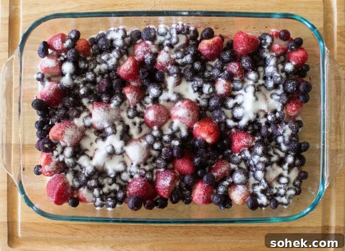 Viral Berry Dump Cake Recipe: Quick No-Fuss Dessert PMID 8 A glass baking dish filled with mixed berries, including strawberries, blueberries, and blackberries, topped with granulated sugar, sits on a wooden surface.