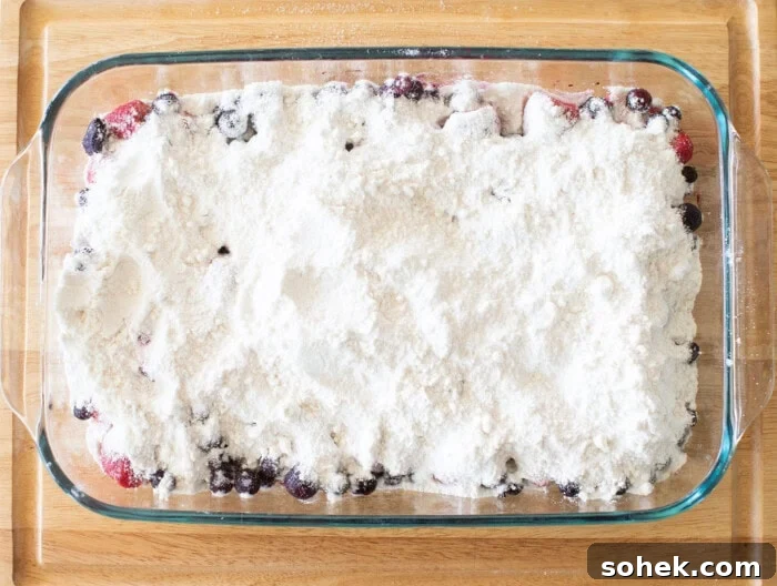 Viral Berry Dump Cake Recipe: Quick No-Fuss Dessert PMID 9 A glass baking dish on a wooden surface filled with a dessert mixture. Visible are mixed berries covered with a layer of flour and sugar, preparing for baking.