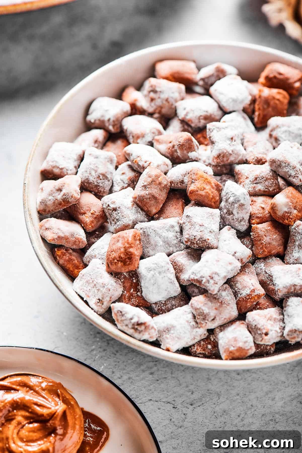 Chocolate Peanut Butter Muddy Buddies Recipe 8 Close-up shot of puppy chow in a white bowl, to show the texture. Some pieces are fully coated in powdered sugar, while others are not.