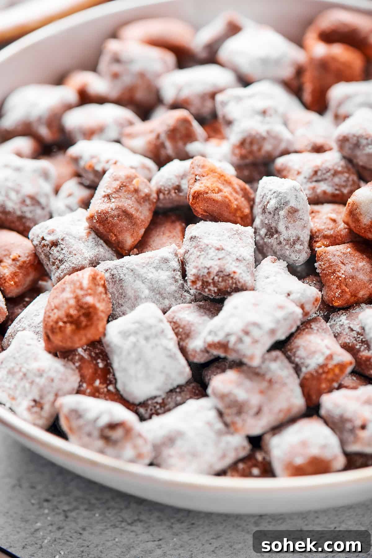 Who’s up for Muddy Buddies? Smooth chocolate, salty peanut butter, and a light dusting of powdered sugar make this crispy Chex cereal snack a true favorite! Close-up shot of puppy chow in a white bowl, to show the texture. Some pieces are fully coated in powdered sugar, while others are not.