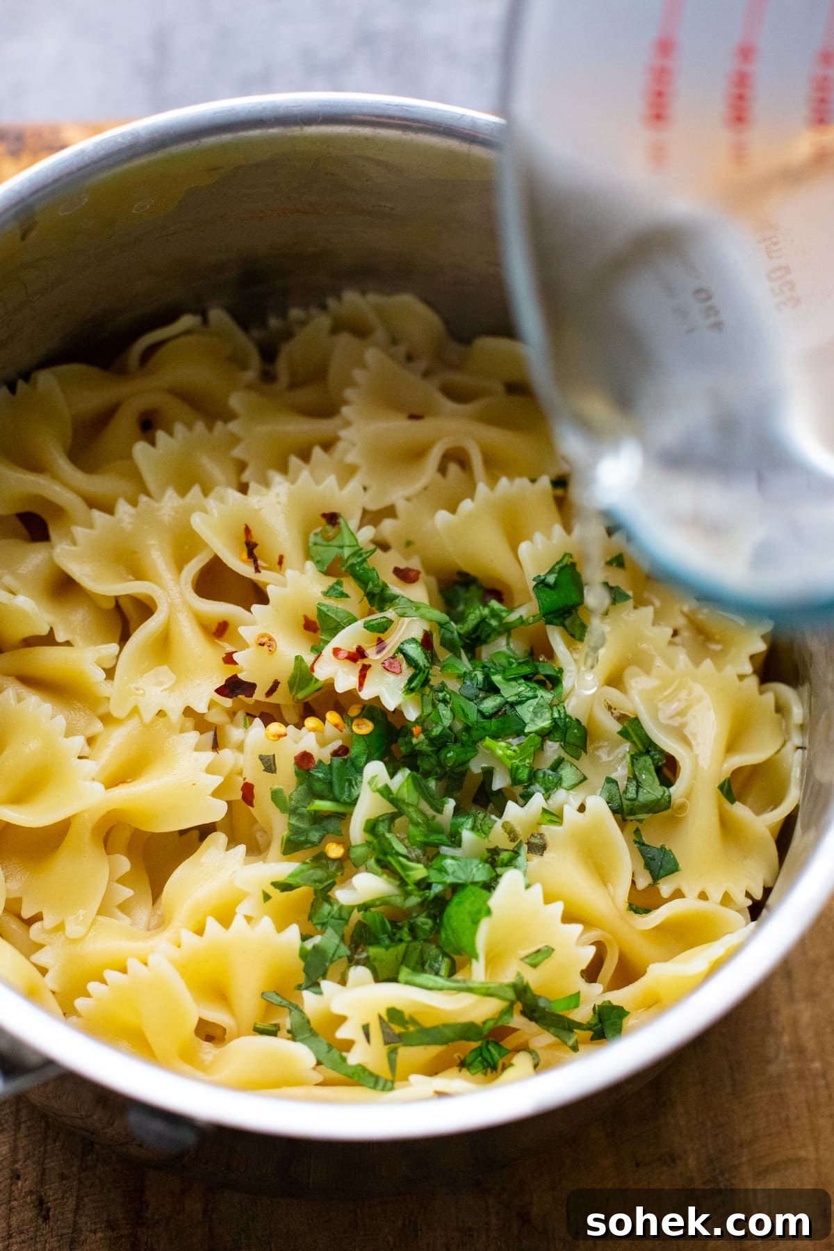 water being poured into pot with farfalle pasta and chopped herbs.