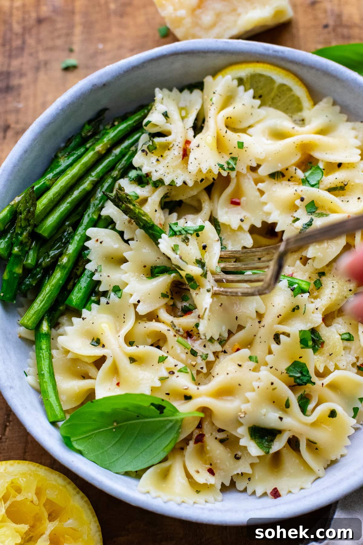 fork pricking bowtie pasta noodles and asparagus in a white ceramic bowl on a brown wooden board.