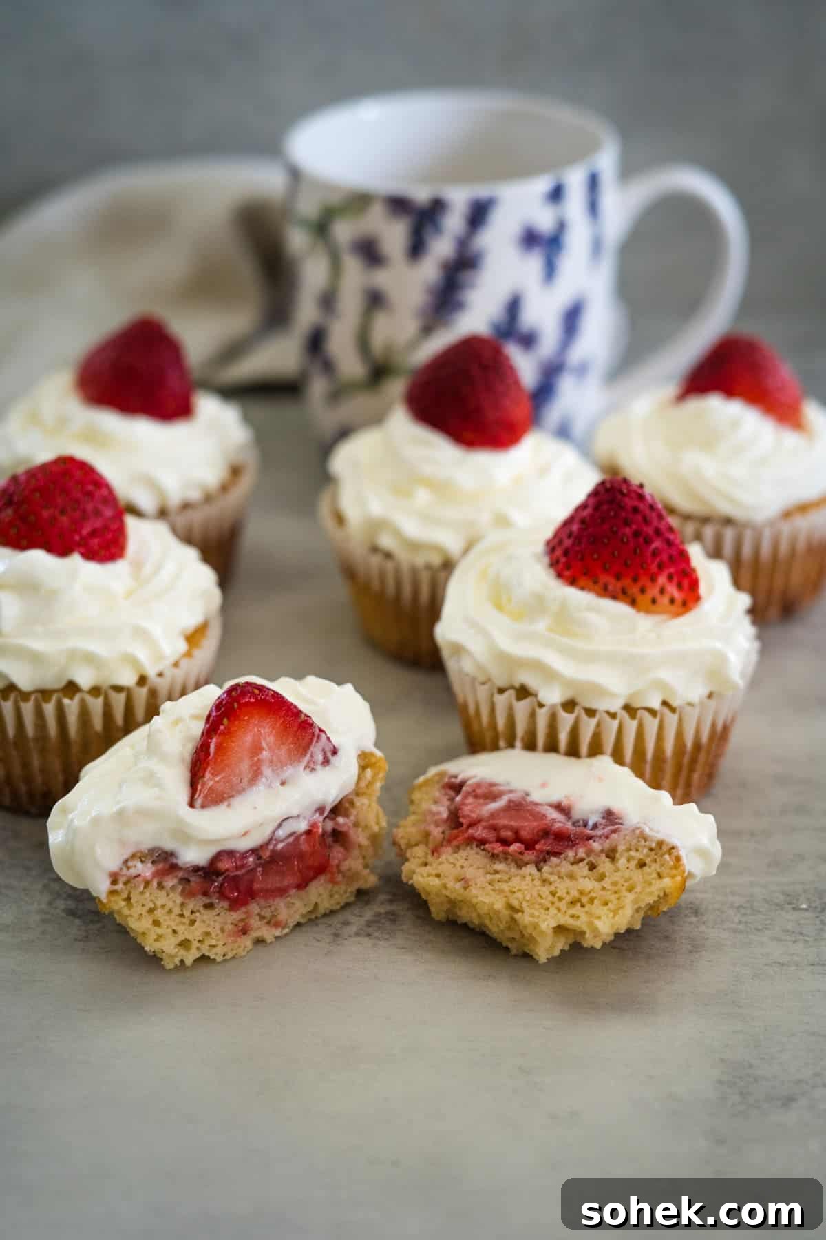 Strawberry Cream Cupcakes: Moist Vanilla Cupcakes with Fresh Strawberry Frosting 2 Several strawberry cream cupcakes topped with whipped cream and strawberries, one of which is cut in half to show the strawberry filling. A floral cup is in the background.
