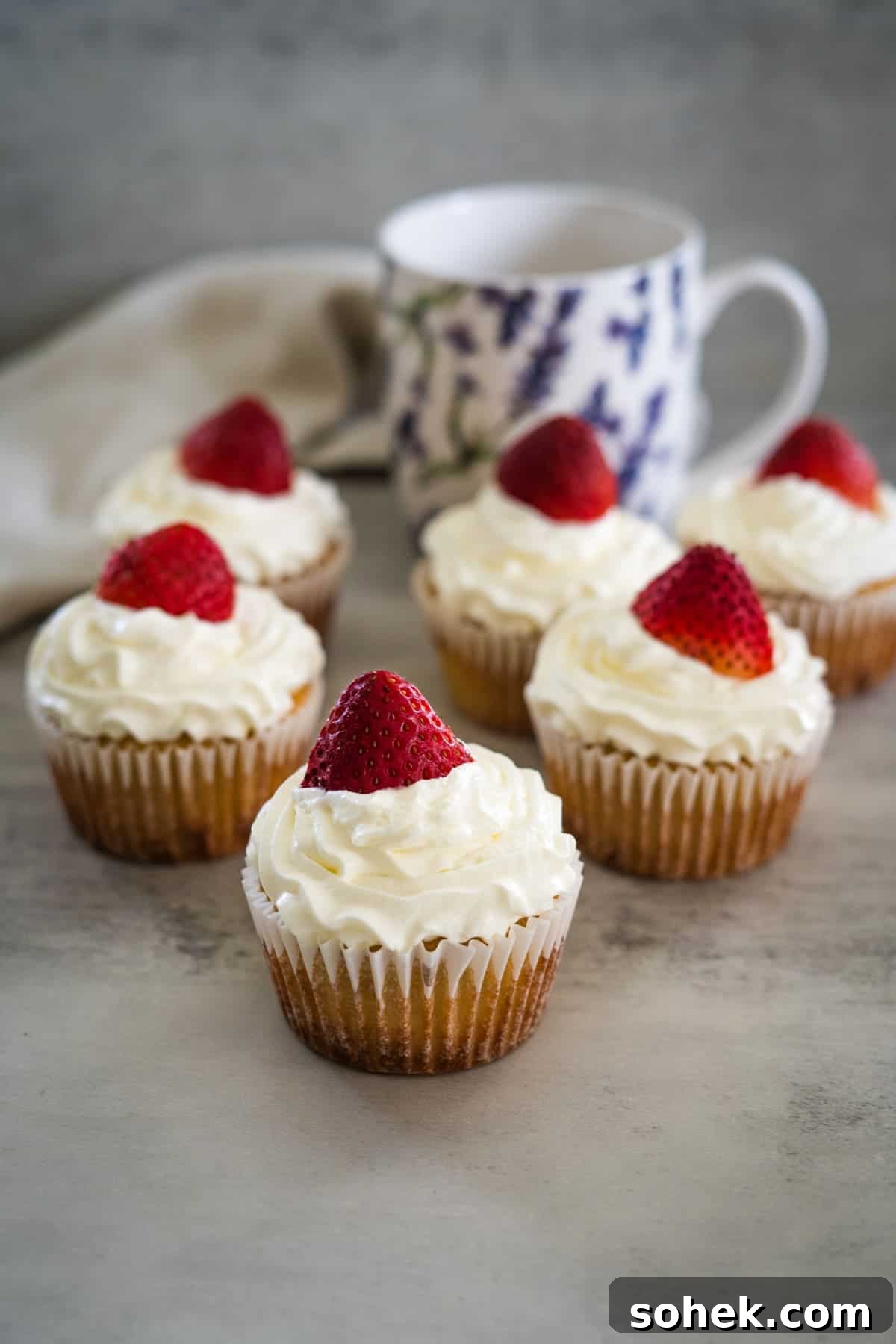 Strawberry Cream Cupcakes: Moist Vanilla Cupcakes with Fresh Strawberry Frosting 5 Six strawberry cream cupcakes topped with white frosting and a strawberry, arranged on a grey surface with a floral mug in the background.