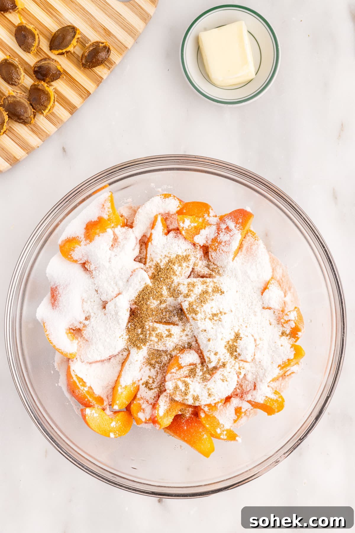 Glass bowl of sliced apricots with sugar, flour, and coriander sprinkled over, pat of butter and cutting board with apricot pits.