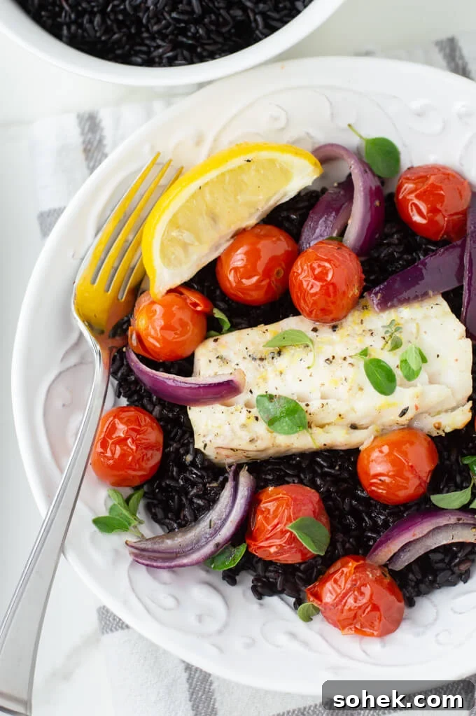 Oven-Baked Cod with Roasted Blistered Tomatoes 2 A white dish with baked cod and cherry tomatoes over black rice. A lemon wedge sits next to it.