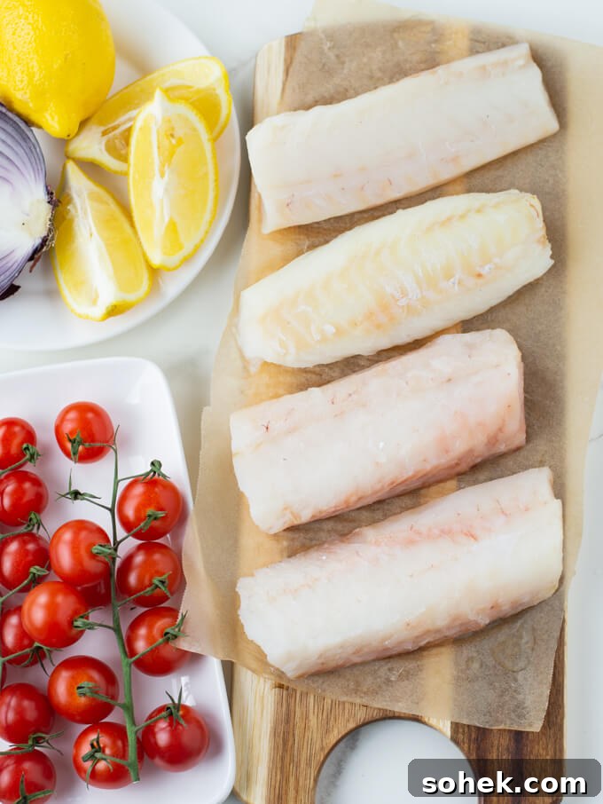 Oven-Baked Cod with Roasted Blistered Tomatoes 3 Thawed Cod fillets sit on parchment paper on a wooden board. Cherry tomatoes and lemon wedges sit next to it.