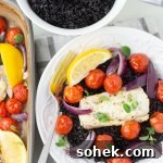 A white dish with baked cod and cherry tomatoes over black rice with a lemon wedge. The baking pan of cod and a bowl of black rice sit next to the dish.
