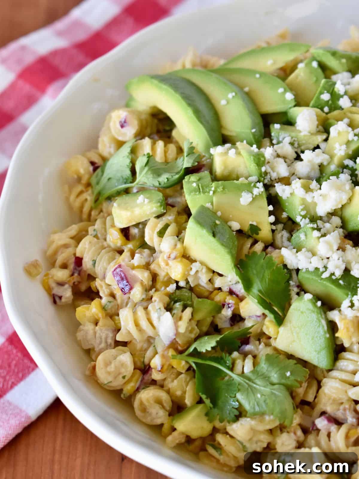Mexican Elote Pasta Salad: Creamy Street Corn Pasta Recipe 8 Elote Pasta Salad in a white bowl with avocado on top and a checkered napkin in the background.