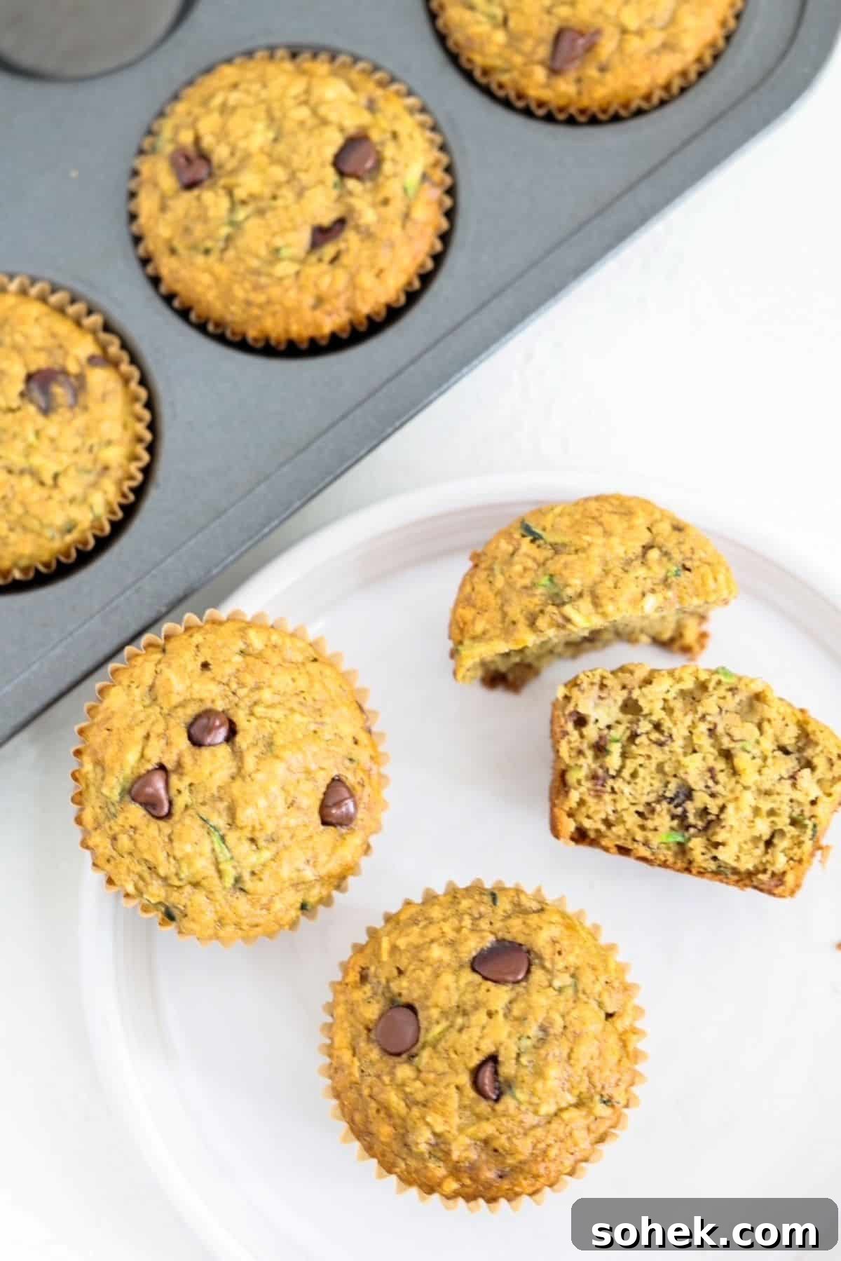 Banana Zucchini Chocolate Chip Muffins Recipe 2 Two whole and two halves of banana chocolate chip muffins on a whit plate with a muffin tin of muffins behind it.