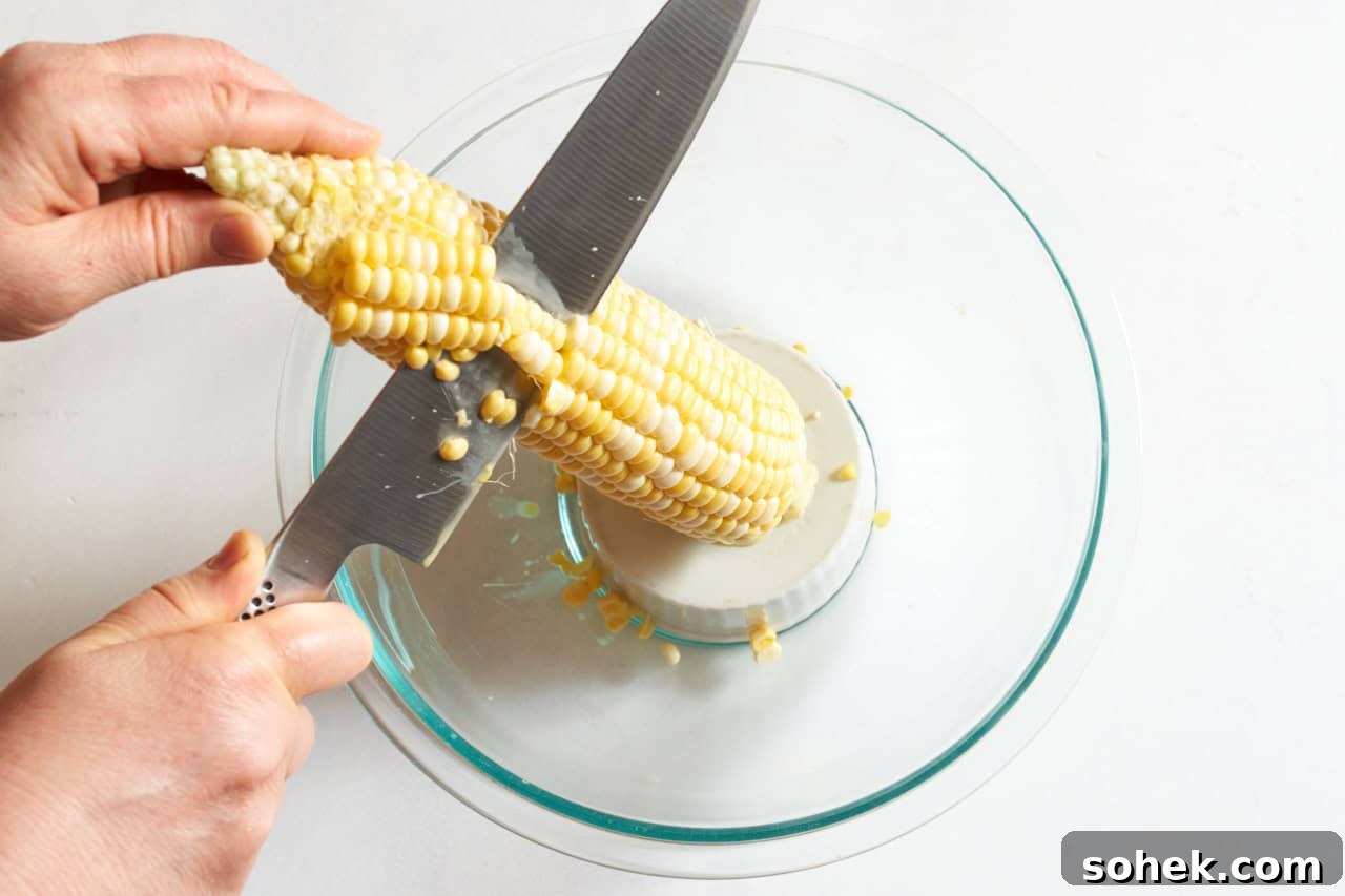 A woman's hands shown cutting corn off the cob with a chef's knife by standing it up on a small bowl set inside a larger one.
