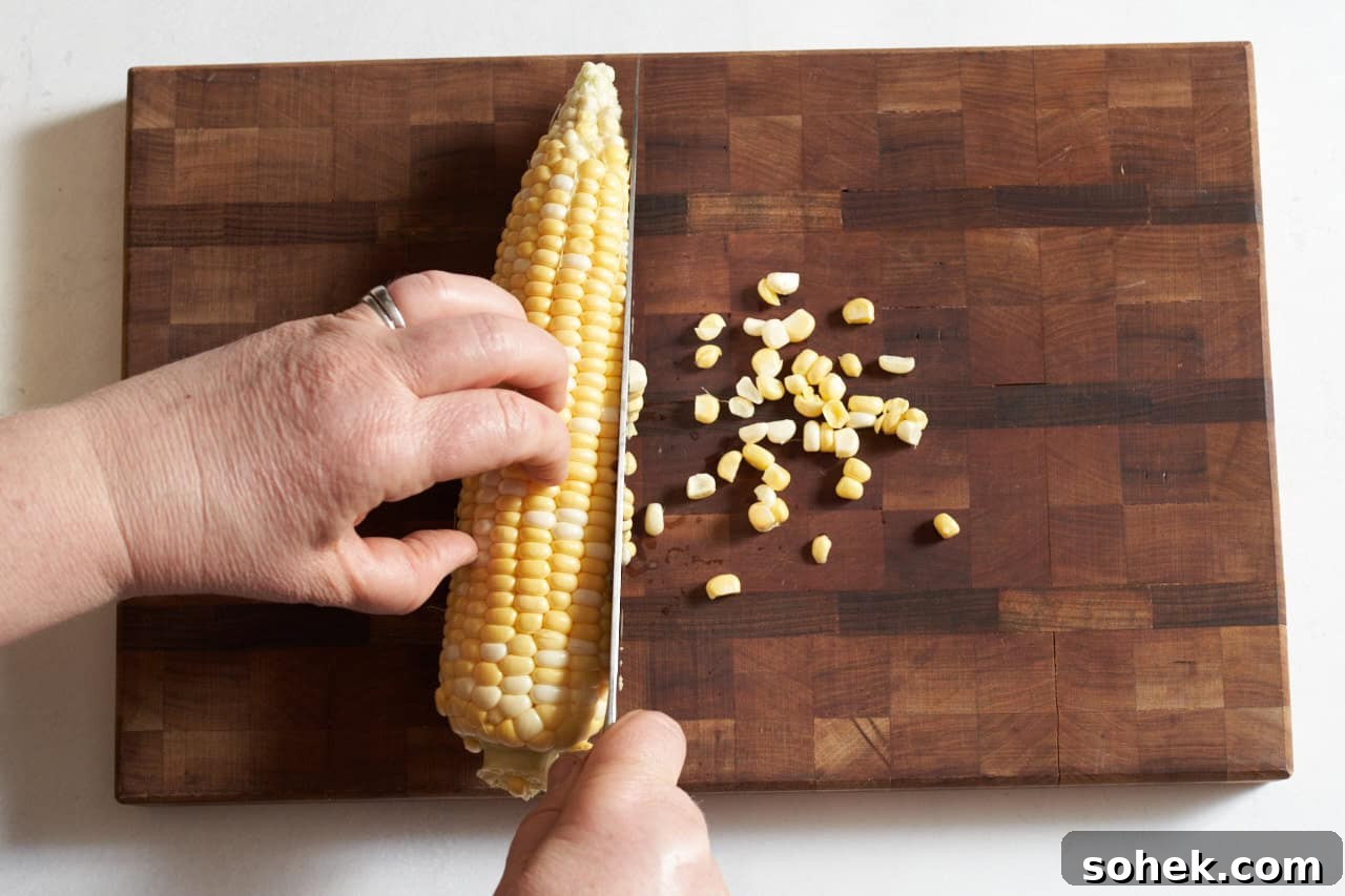 A woman's hands shown cutting corn off the cob with a chef's knife by laying it flat on a wooden cutting board.