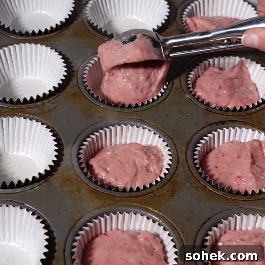 Strawberry Crunch Cupcakes Recipe with Crispy Topping 10 Portioning strawberry crunch cupcake batter into white liners in a muffin tin, preparation step for baking.