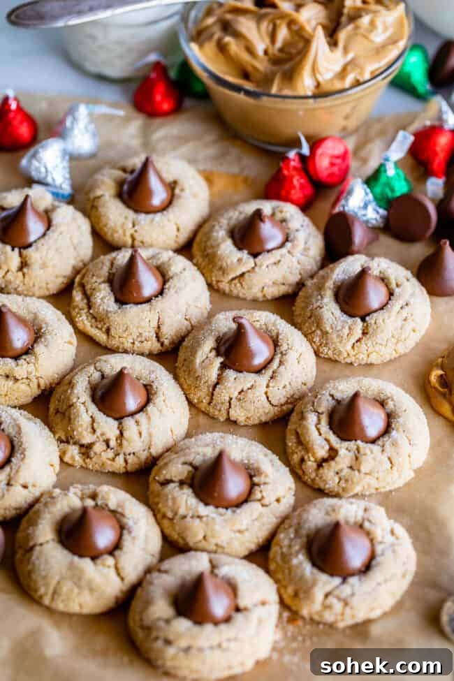 peanut butter blossoms on parchment paper with Hershey's kisses and a bowl of peanut butter in the background.