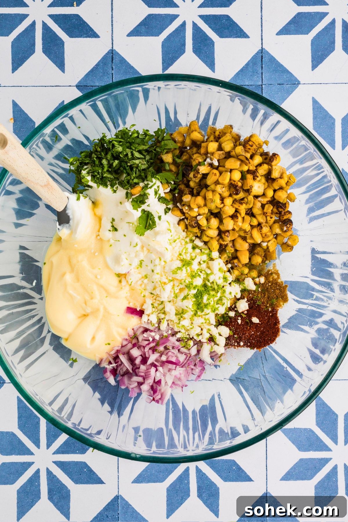Spices, cheese, roasted corn, and other ingredients mixed together in a clear  mixing bowl, to make a corn dip. 