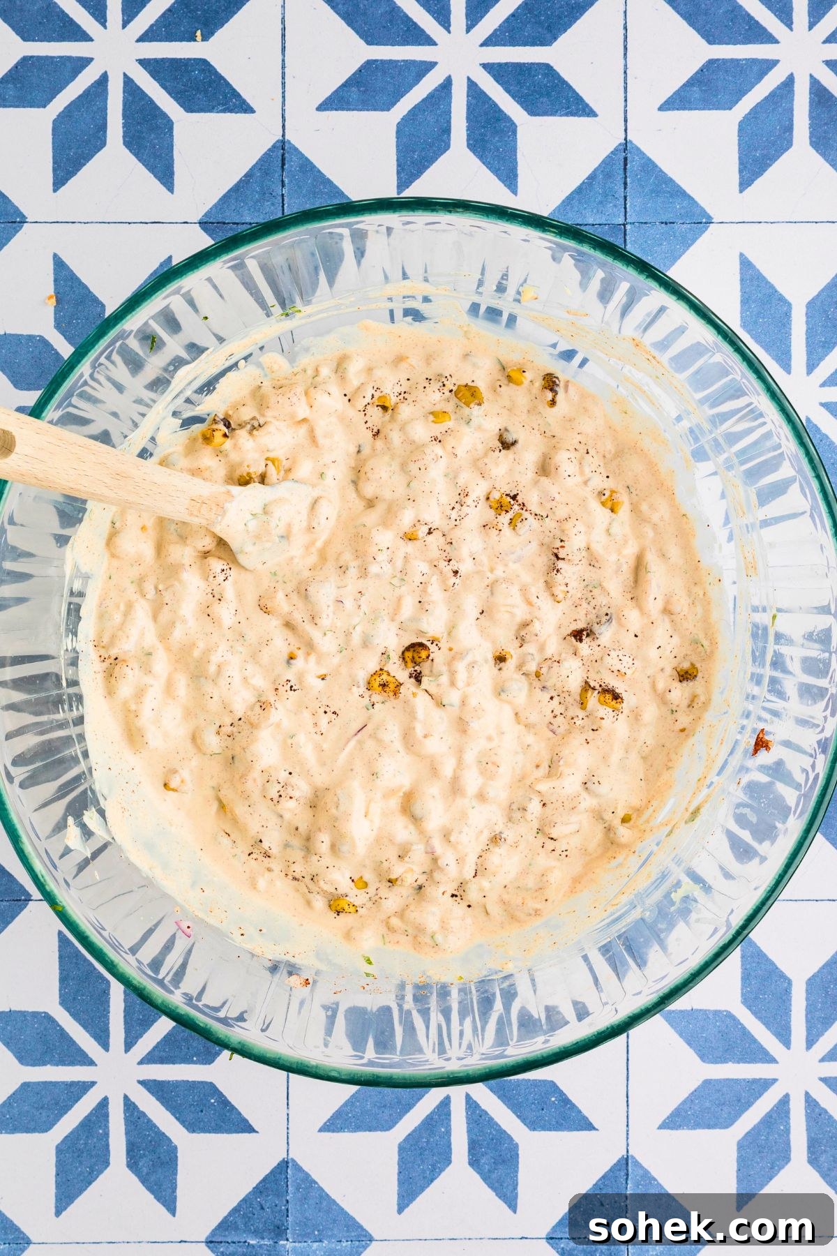 Corn dip ingredients mixed together in a clear glass bowl on a blue tiled table. 