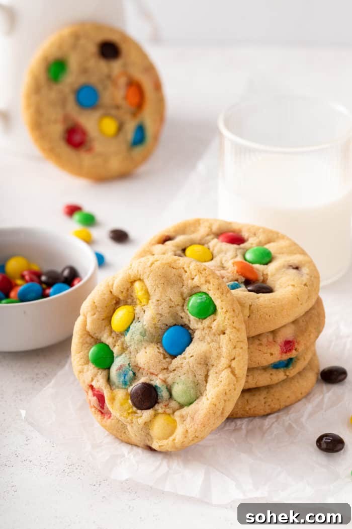 Chewy M&M Cookies Recipe for Perfect Chocolate Crumble 2 4 M&M cookies stacked on a piece of parchment paper in front of a glass of milk, with a 5th cookie leaning against the front of the stack.