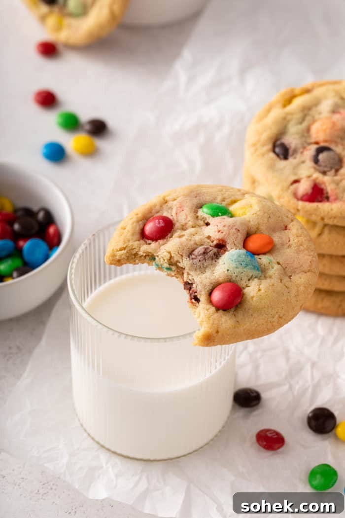 Chewy M&M Cookies Recipe for Perfect Chocolate Crumble 3 M&M cookie with a bite taken out of it set on the edge of a glass of milk.