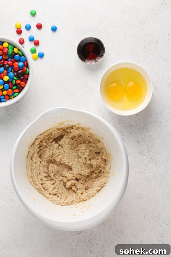 Chewy M&M Cookies Recipe for Perfect Chocolate Crumble 5 Butter and sugar creamed together in a white bowl, set next to smaller bowls of eggs and vanilla.