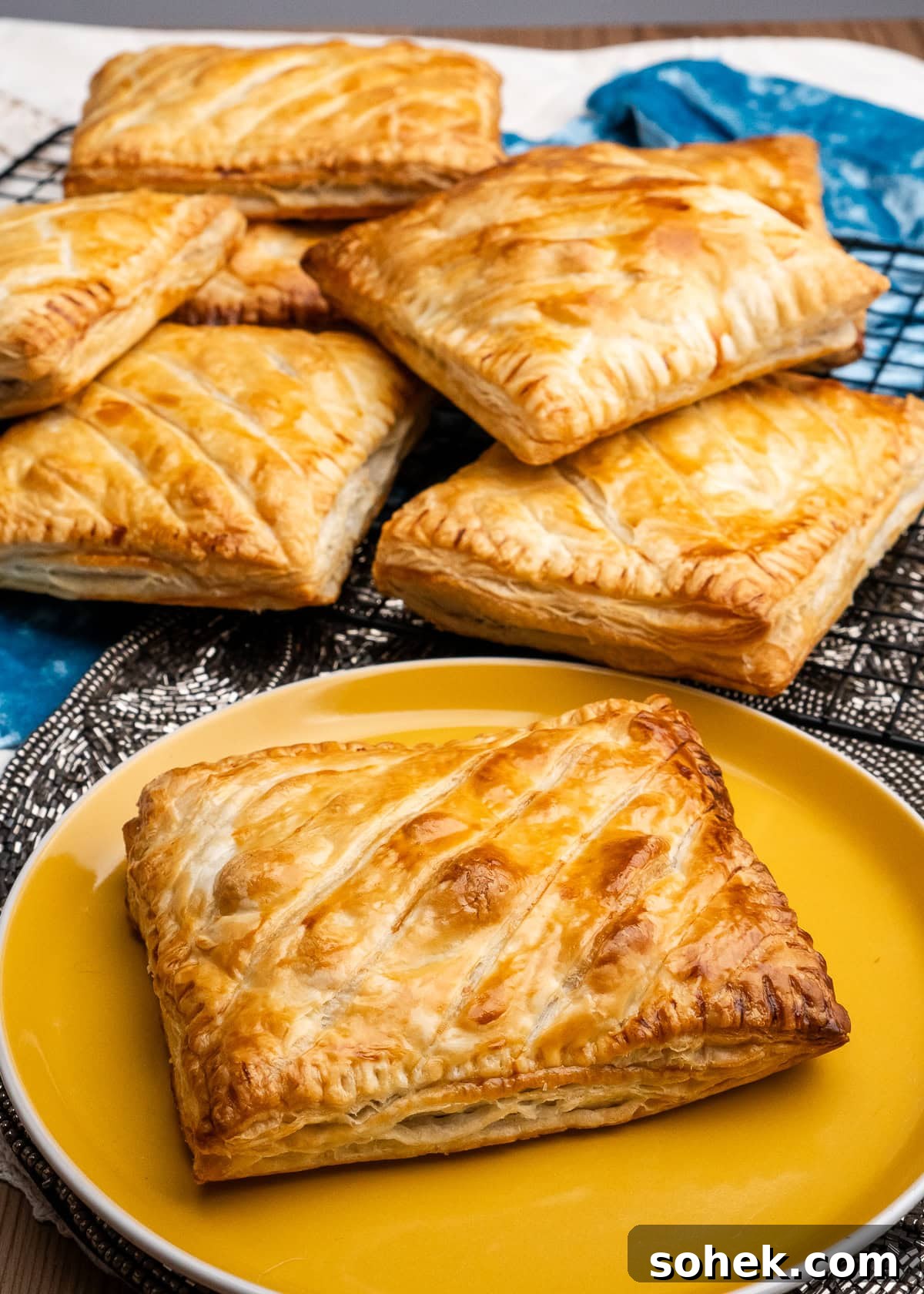 Greggs-Style Steak Bake Recipe: How to Make It at Home 43 A steak bake on a yellow plate and steak bakes stacked up on top of each other in the background on a wire rack with a blue and white cloth under the wire rack