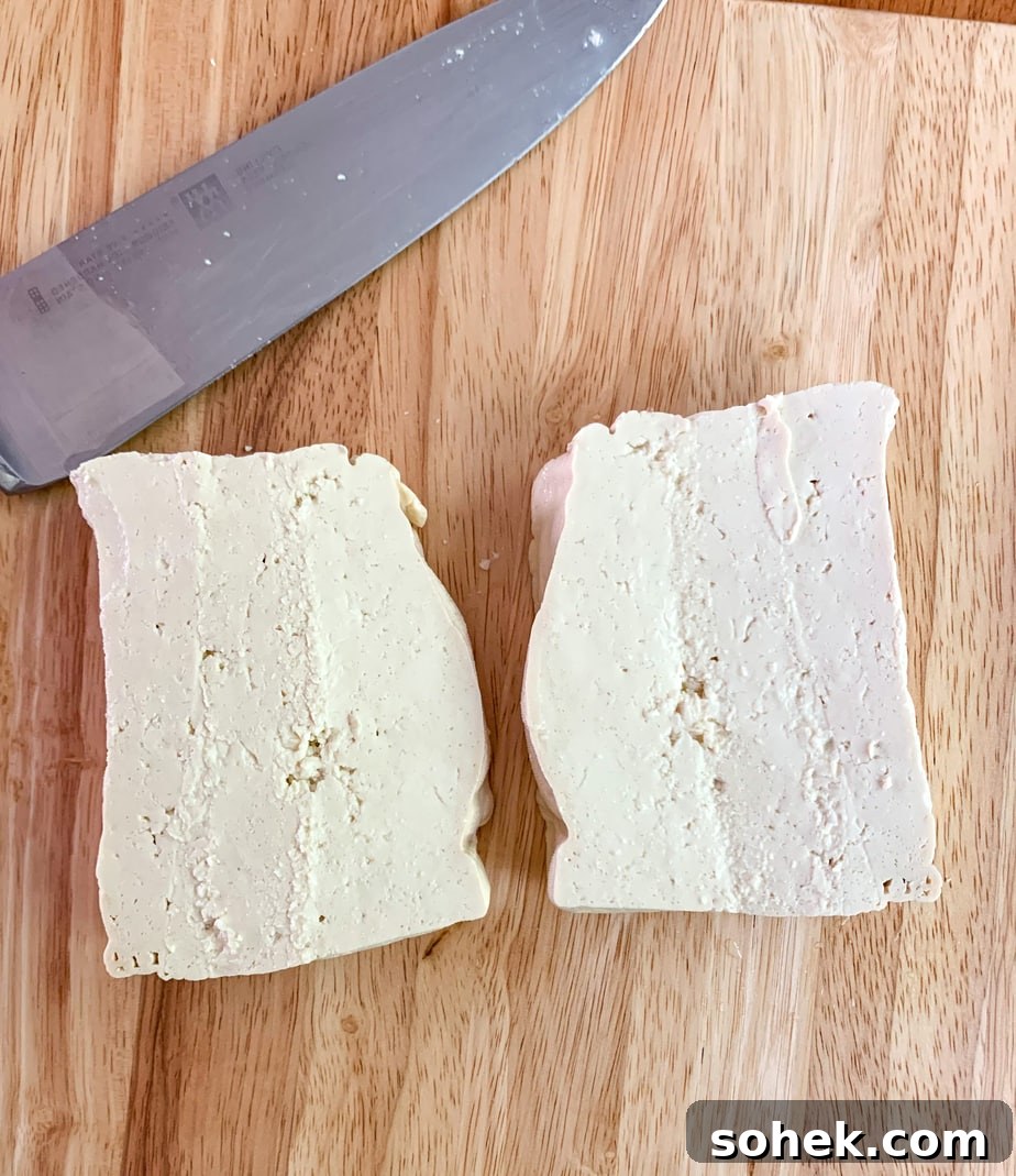A block of tofu sliced in two pieces with a knife, displayed on a butcher block board.
