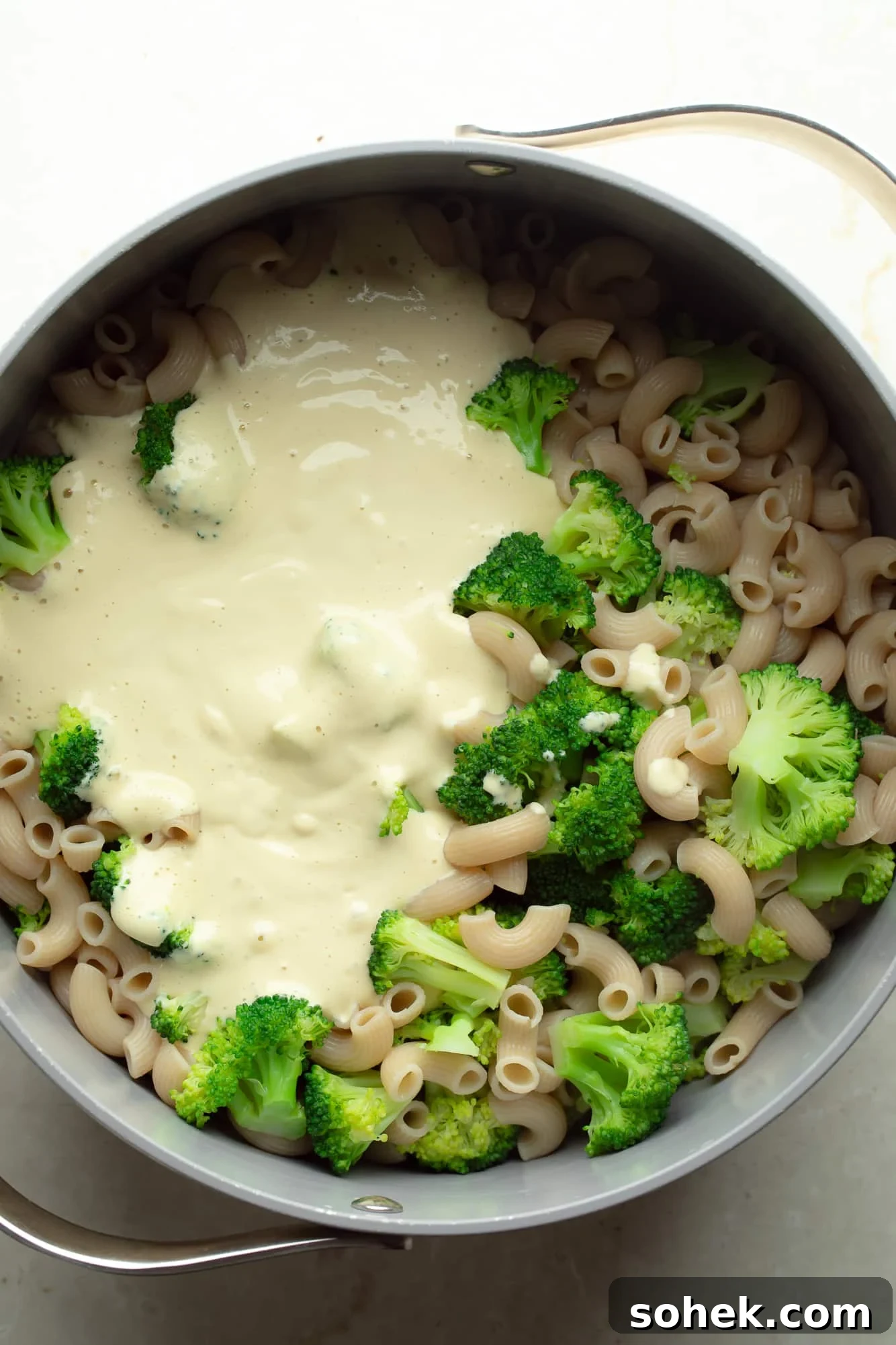 High-Protein Sharp White Mac and Cheese with Broccoli 6 overhead view of white vegan cheese sauce on top of cooked broccoli and pasta in a large pot.