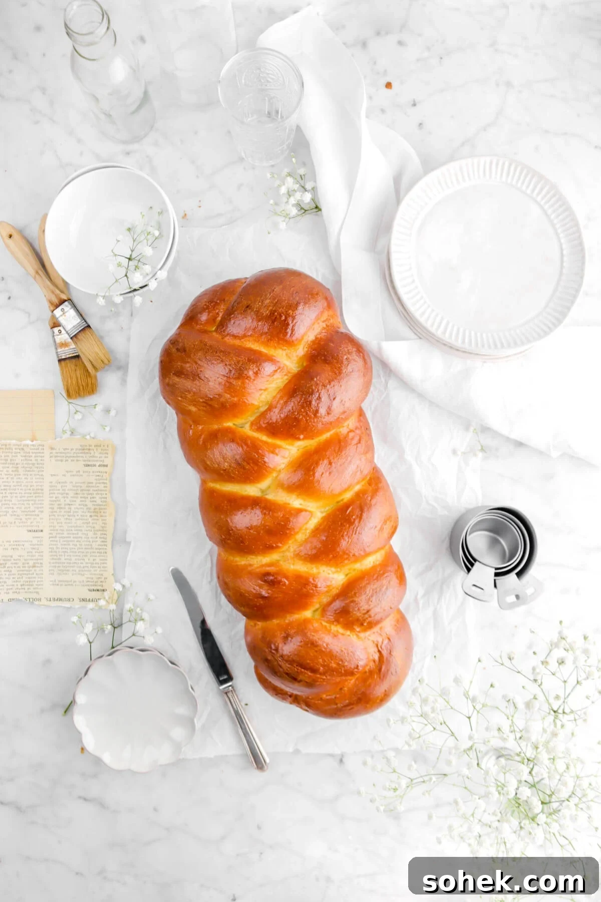 pulled back shot of challah on marble surface with plates, flowers, and a knife
