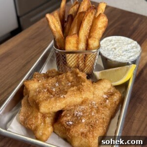 A serving tray with golden brown fish and chips, accompanied by a lemon wedge and a small cup of tartar sauce. The chips are held in a small metal basket, and everything is placed on a wooden table.