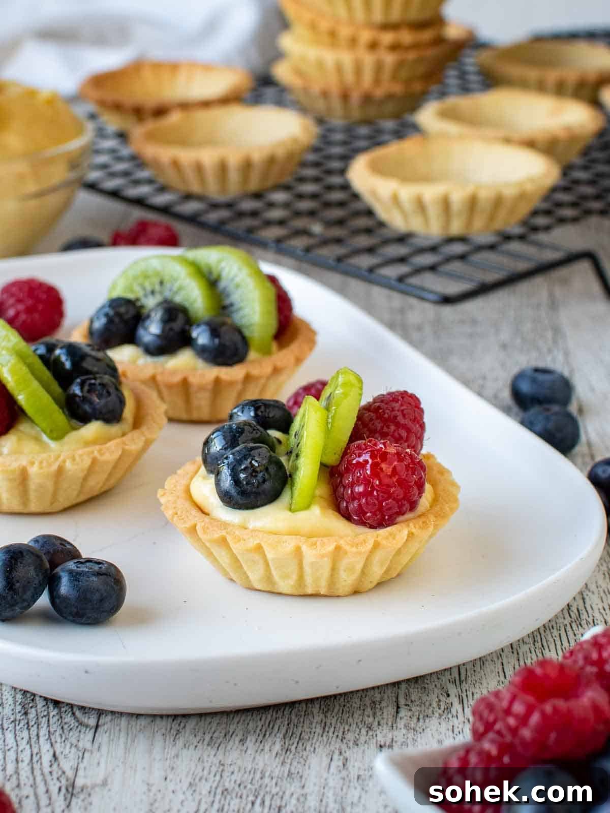 Mini Fresh Fruit Tarts Recipe: Bite-Sized Dessert for Parties 2 Mini fruit tarts on a white oblong plate with tartlet bases in the background.
