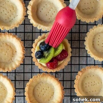 Mini Fresh Fruit Tarts Recipe: Bite-Sized Dessert for Parties 13 Individual fruit tarts filled and topped with fruit being brushed with a red pastry brush.