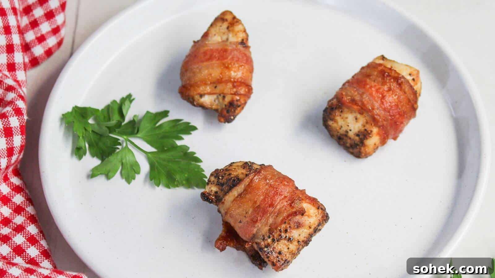 26 Irresistible Air Fryer Game Day Snacks to Serve Guests 13 Three bacon-wrapped pieces of chicken on a white plate with a sprig of parsley and a red and white checkered cloth on the side.