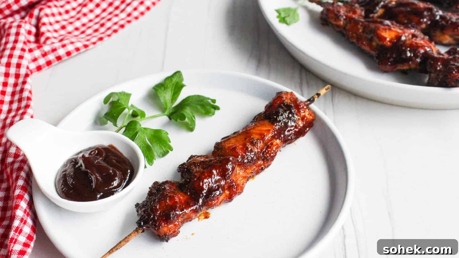 26 Irresistible Air Fryer Game Day Snacks to Serve Guests 15 A single barbecued chicken skewer on a white plate, with a small bowl of barbecue sauce and a parsley garnish, next to a plate of additional chicken skewers.