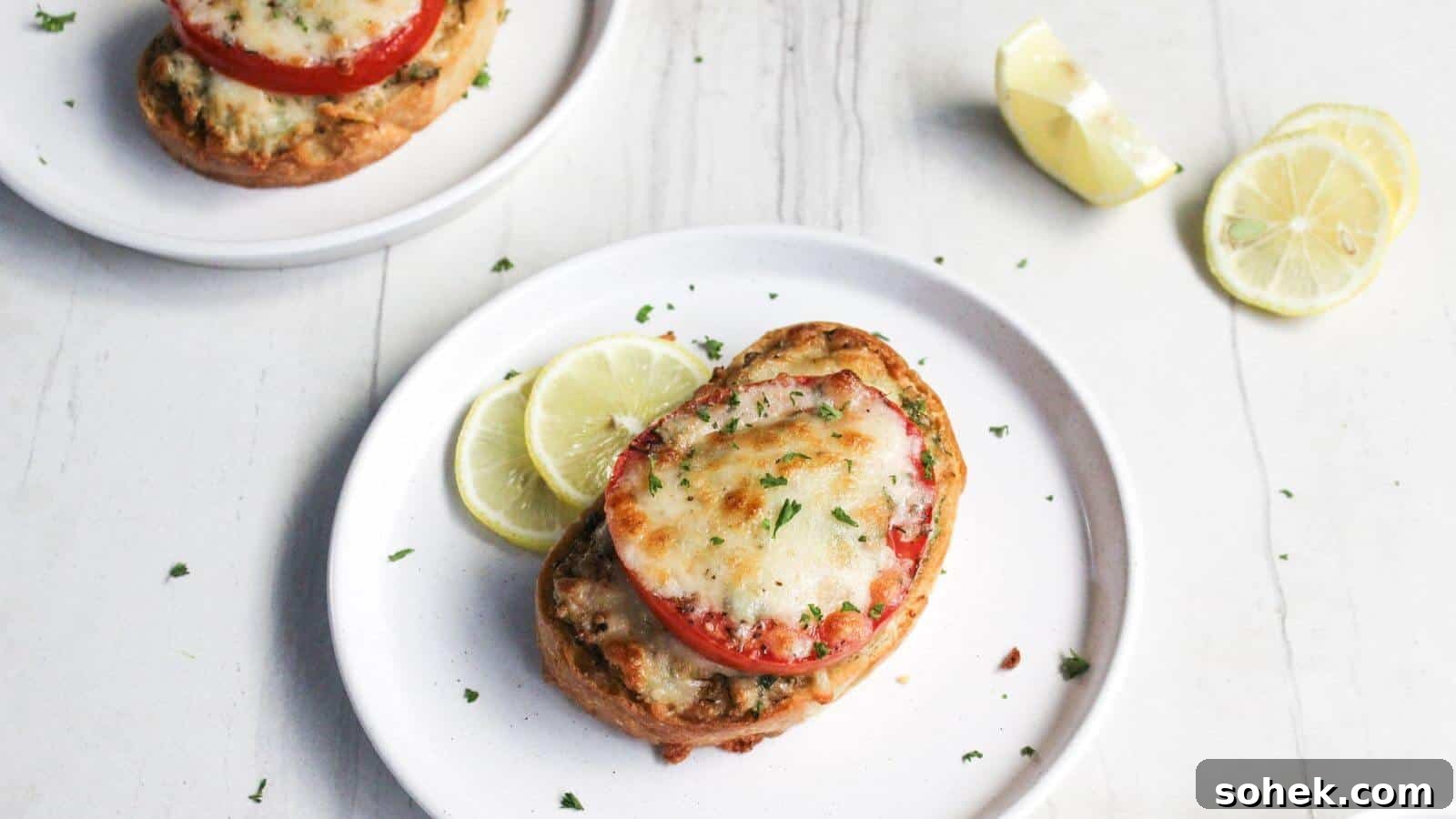 26 Irresistible Air Fryer Game Day Snacks to Serve Guests 19 Two plates of crab melt with sliced tomatoes and melted cheese, garnished with lemon slices on a white wooden surface.