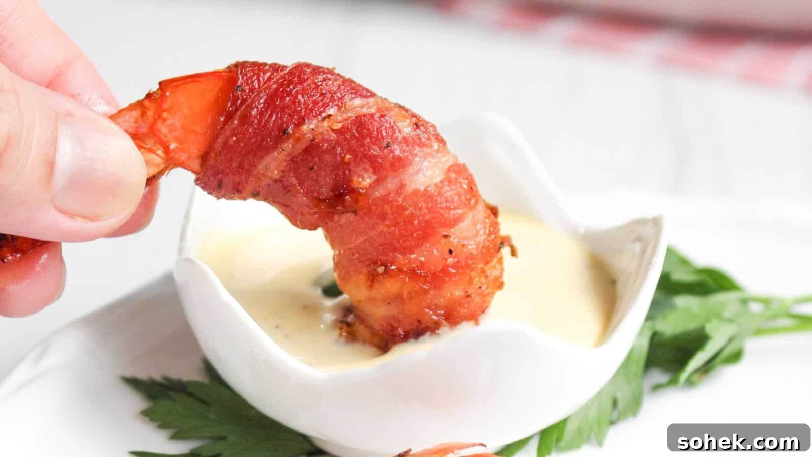 26 Irresistible Air Fryer Game Day Snacks to Serve Guests 8 A hand holding a bacon-wrapped shrimp dipped in a small bowl of sauce, with a green leaf garnish on the side.