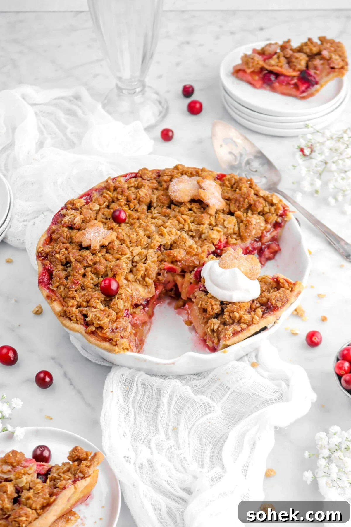 Apple Cranberry Crumble Pie Recipe with Buttery Oat Topping 5 angled front shot of apple cranberry pie with two slices beside, on cheesecloth, with cranberries and a pie plate around.