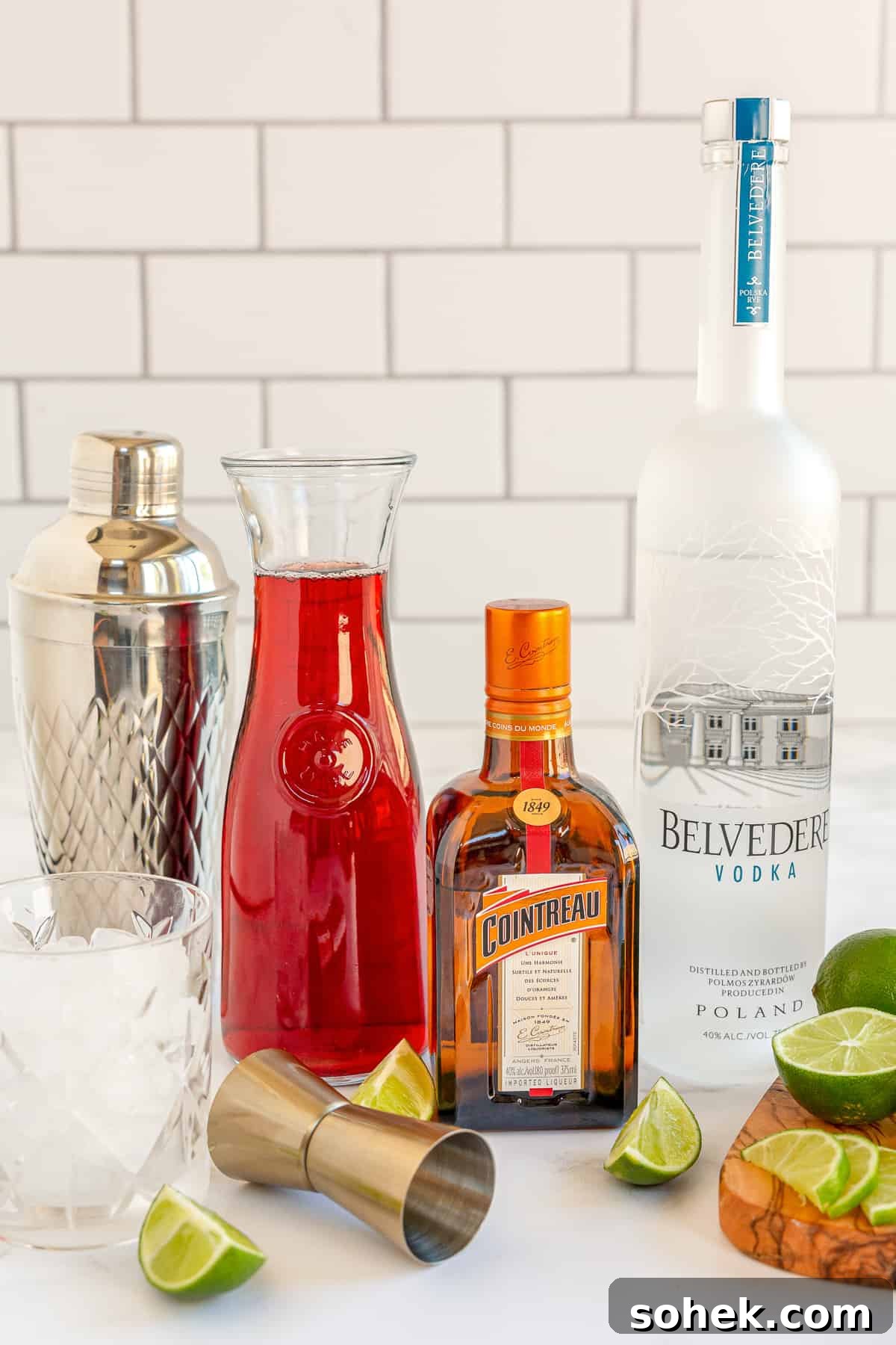 Classic Cosmopolitan Cocktail Recipe: How to Mix a Perfect Cosmopolitan 6 A bottle of vodka, Countreau, and a carafe of cranberry juice surrounded by a cocktail shaker and lime wedges.