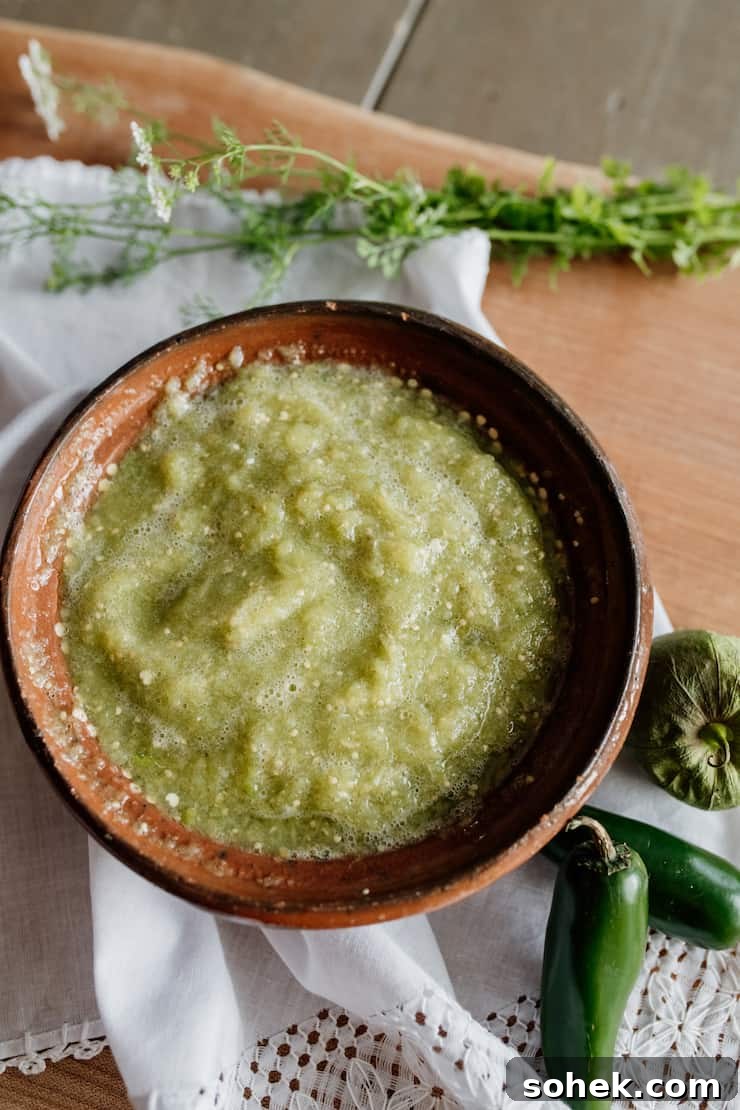 salsa verde in a brown Mexican pottery bowl on a white cloth side of two jalapeño peppers and a tomatillo. 