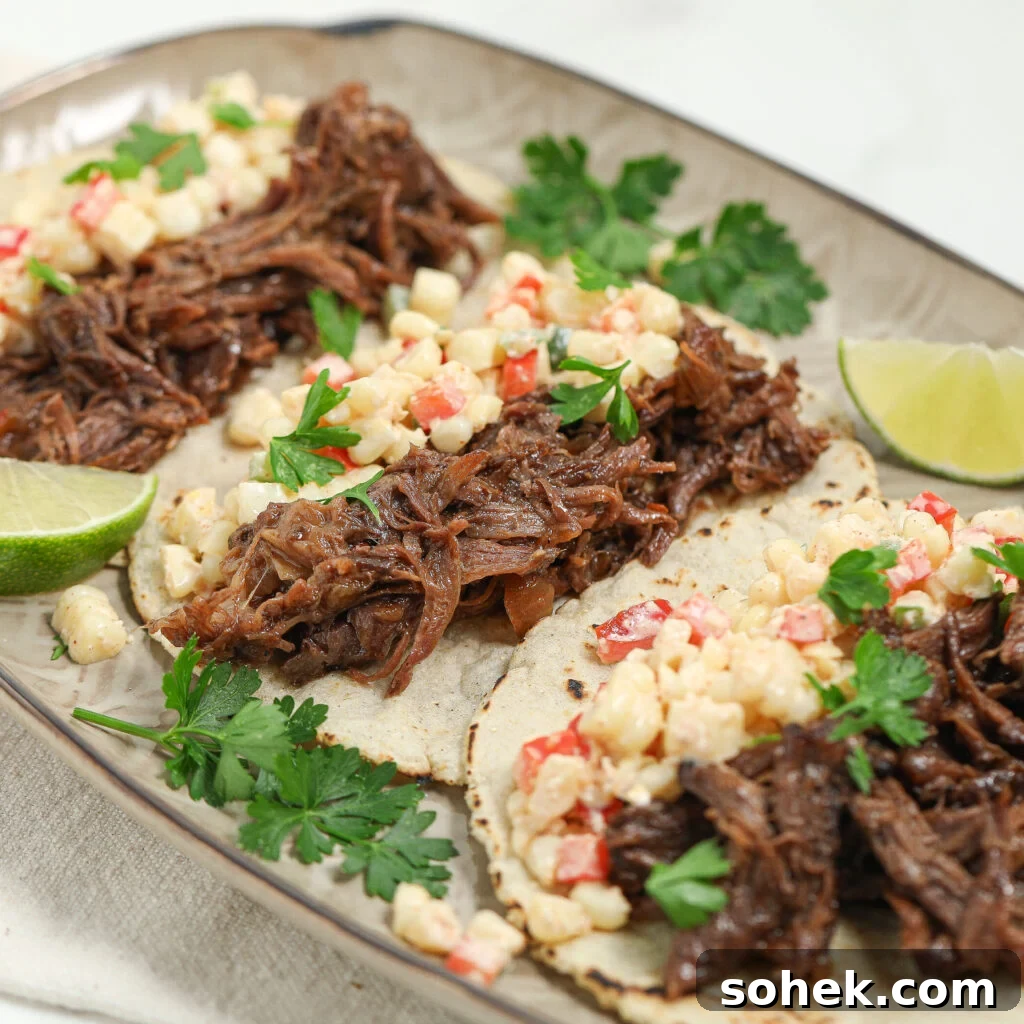 Chuck Roast Tacos with Mexican Street Corn Salsa (Street-Style) 3 Three corn tortillas topped with shredded beef, a corn and red pepper salsa, and fresh cilantro are arranged on a plate with lime wedges and parsley garnish.